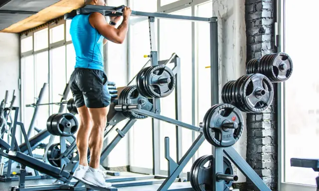 Hombre entrenando en gimnasio, con barra en hombros. Se ven discos de pesas y máquinas.