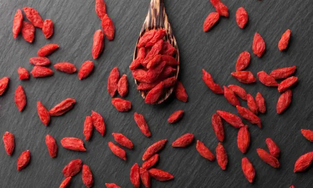 Cucharón de madera lleno de bayas de goji rojas y secas, esparcidas sobre una superficie oscura.