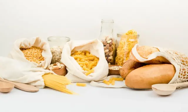Variedad de alimentos para una carga de hidratos: pasta, pan, avena, frutos secos y legumbres en bolsas de tela y frascos.