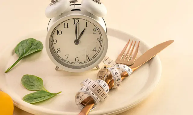 Reloj, plato, cubiertos con cinta métrica y hojas verdes. Símbolos de los beneficios del ayuno intermitente y un estilo de vida saludable.