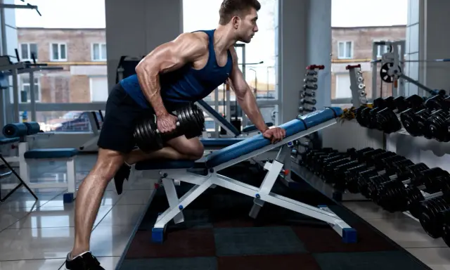Hombre musculoso realiza remo sentado con mancuerna en gimnasio.