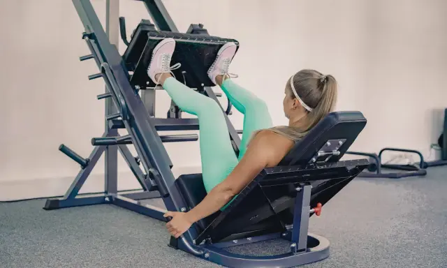 Mujer rubia con leggings turquesa y zapatillas rosas haciendo prensa ejercicio en una máquina de gimnasio.