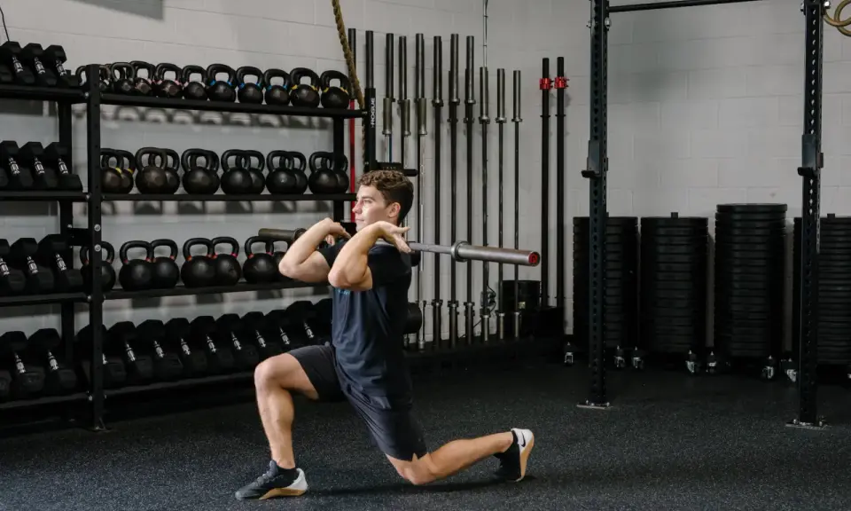 Hombre realizando un front rack lunge con barra en un gimnasio, rodeado de pesas y barras.