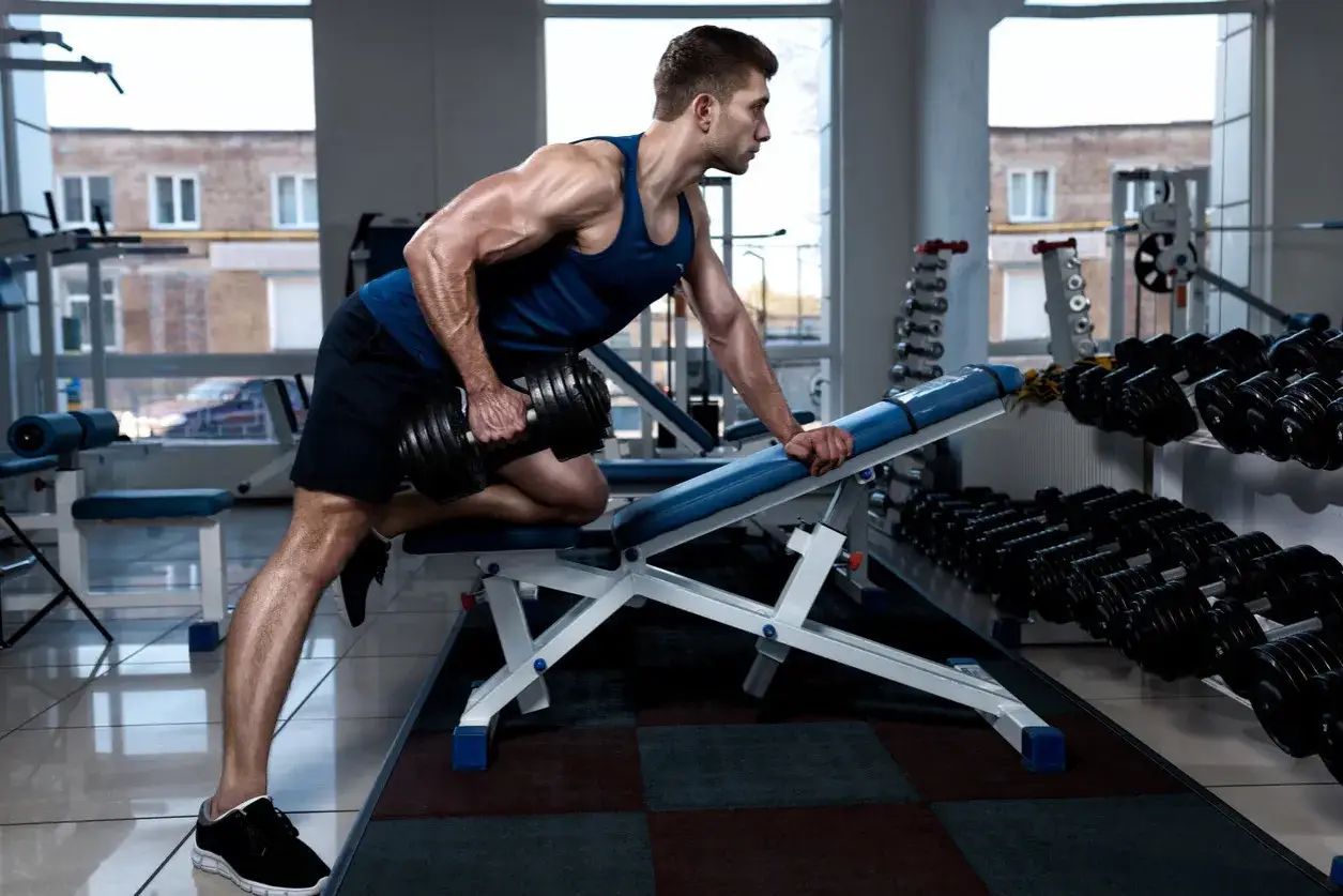 Hombre musculoso realiza remo sentado con mancuerna en gimnasio.