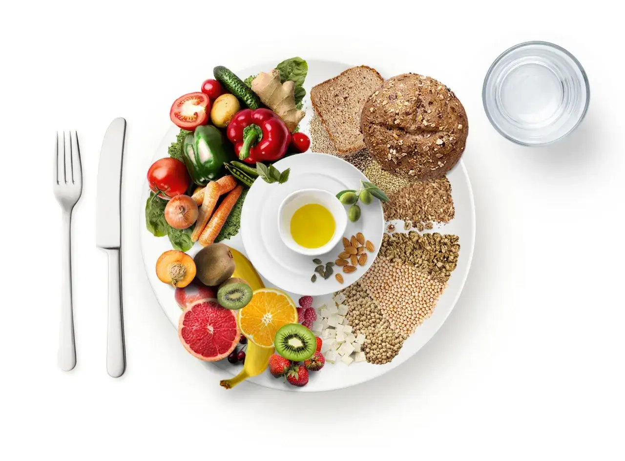 Plato con variedad de frutas, verduras, granos, pan integral, aceite de oliva y agua. Una lista de alimentos que comen los veganos.