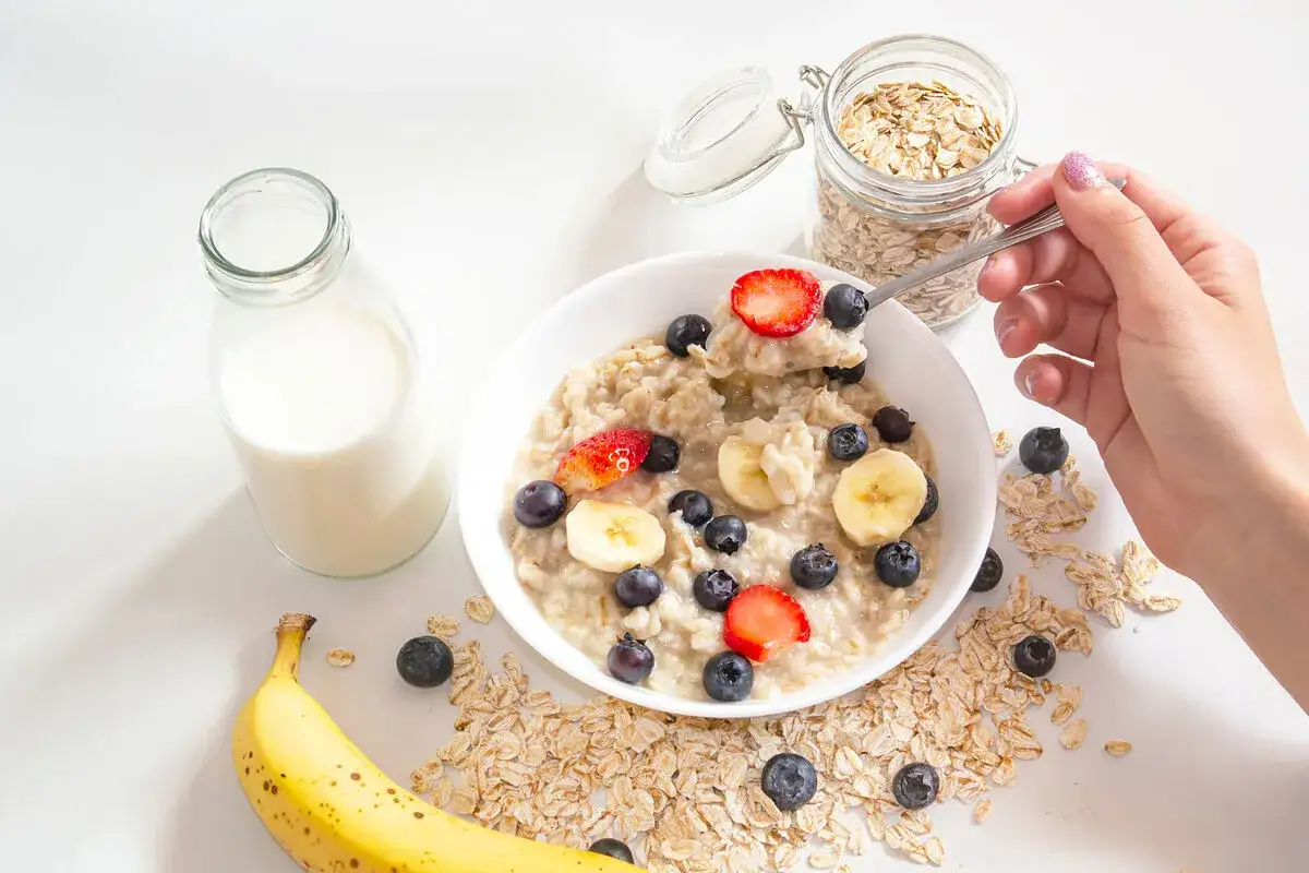 Un taz&oacute;n de avena con fresas, ar&aacute;ndanos y pl&aacute;tano. Una mano toma una cucharada, lista para disfrutar. &iexcl;As&iacute; se come avena!