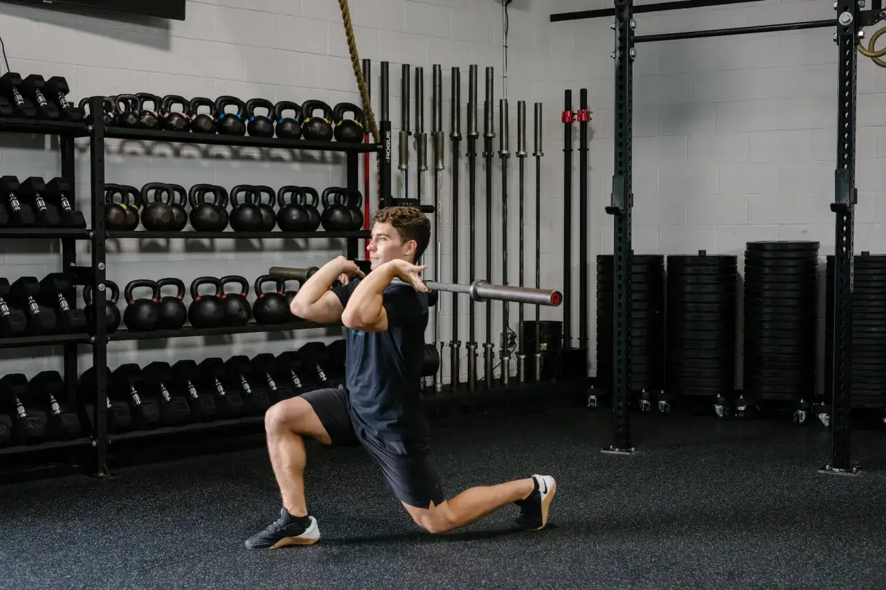 Hombre realizando un front rack lunge con barra en un gimnasio, rodeado de pesas rusas y barras.