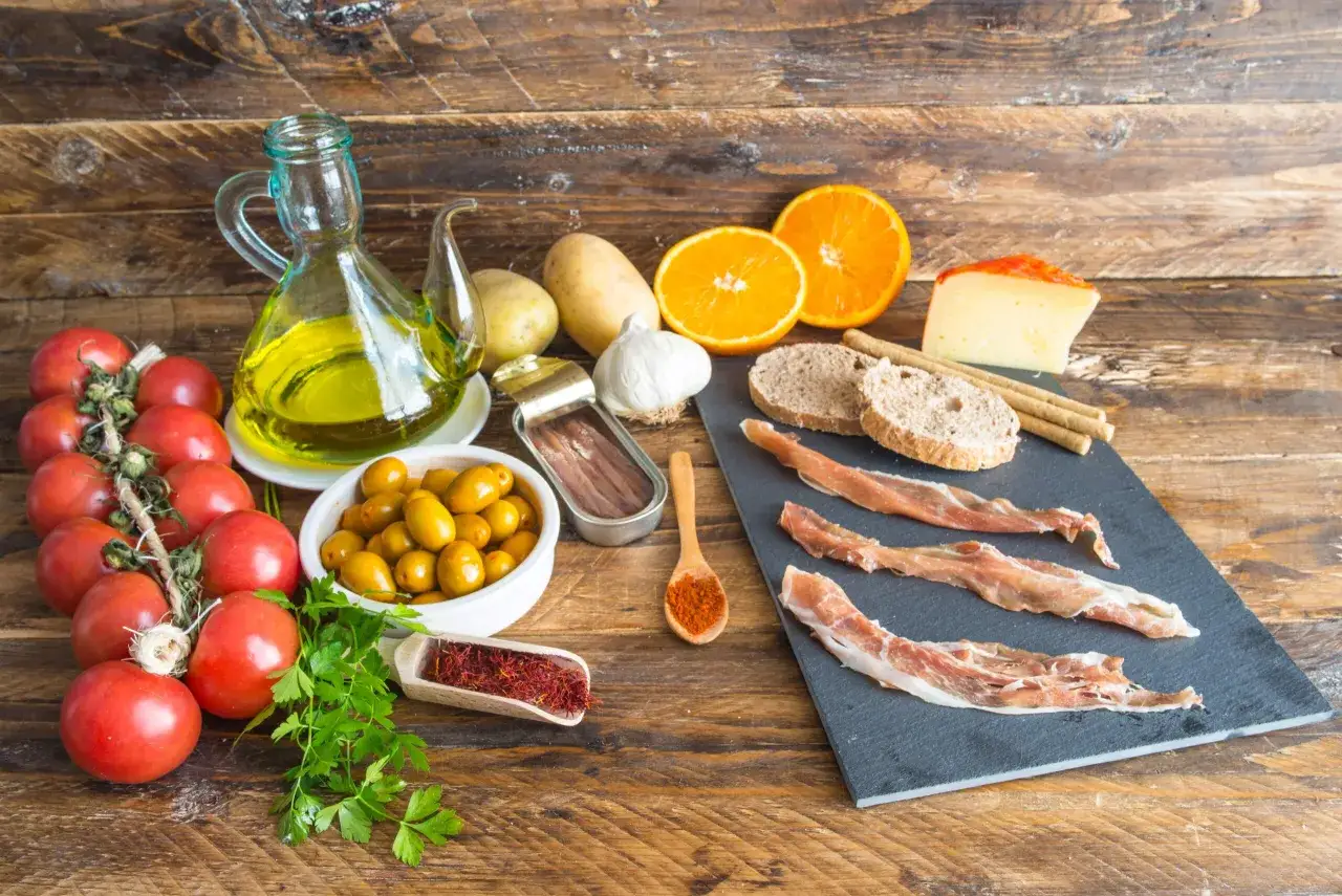Ingredientes para una dieta baja en FODMAP: tomates, aceitunas, jamón serrano, naranjas, patatas, ajo, aceite de oliva y especias.