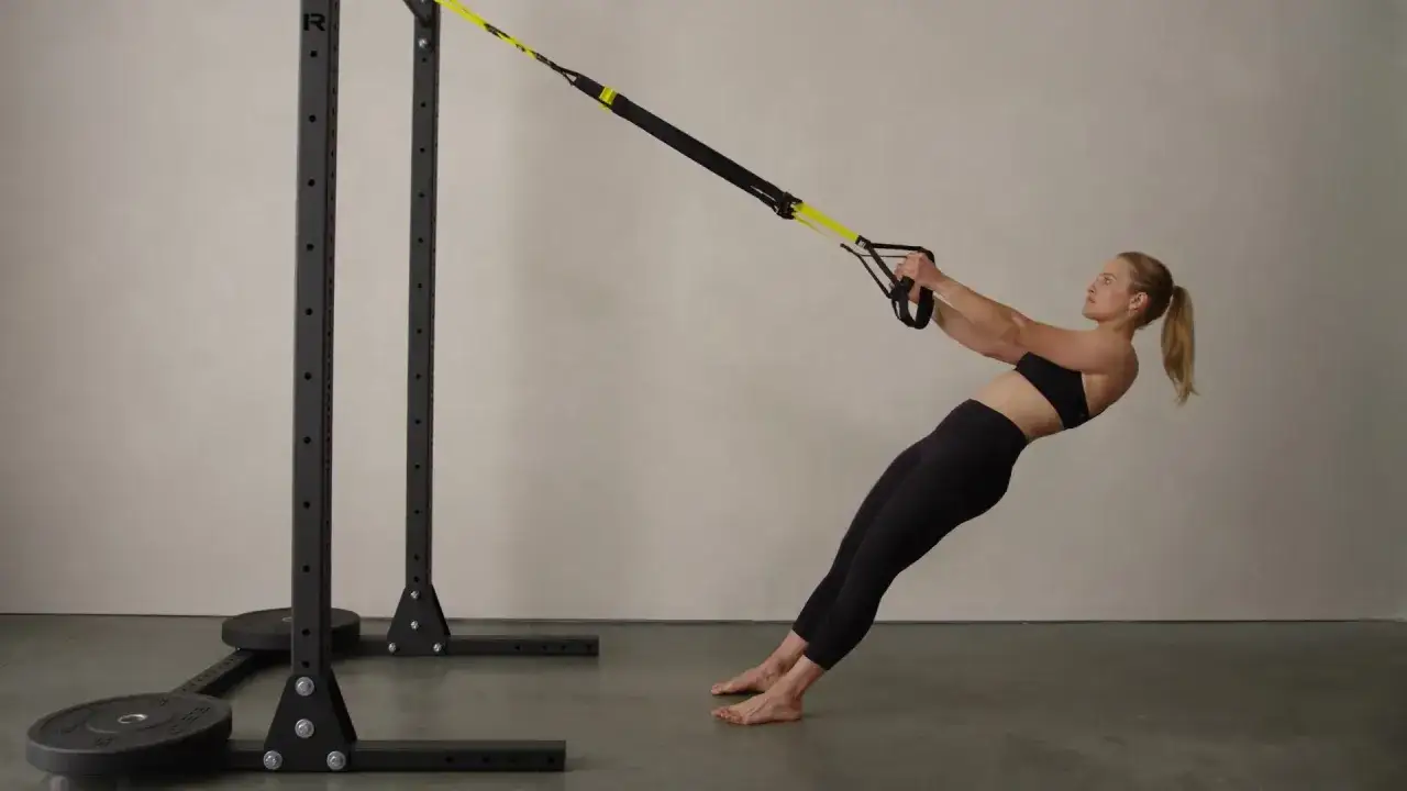 Mujer realizando remo TRX, con el cuerpo inclinado y los pies apoyados en el suelo, usando un equipo de entrenamiento de suspensión.
