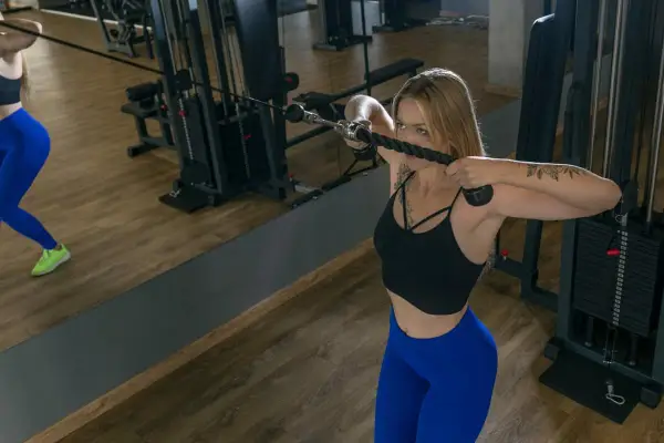 Mujer rubia realiza ejercicio face pull en polea alta, concentrada en su entrenamiento de hombros y espalda.