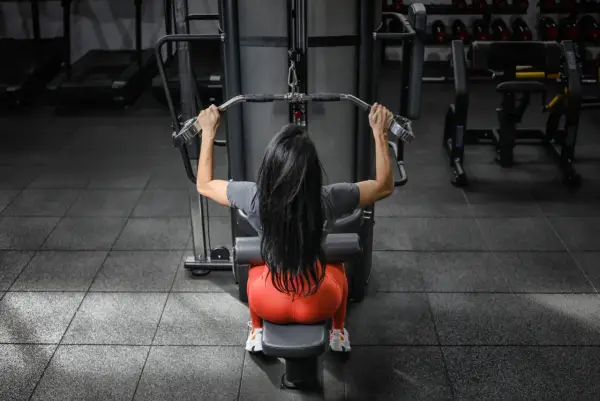 Mujer con cabello largo y negro realiza ejercicios de antebrazo en polea, sentada en una máquina de gimnasio.