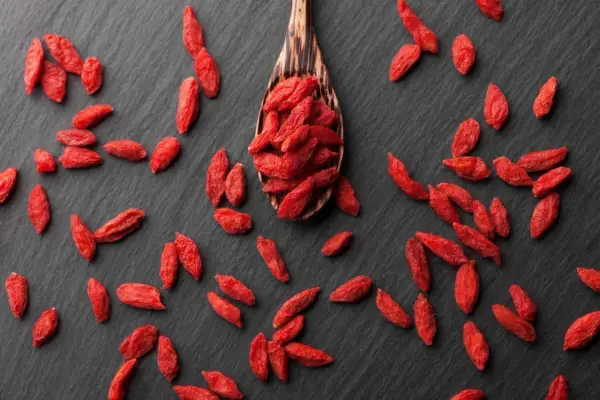 Cucharón de madera lleno de bayas de goji rojas y secas, esparcidas sobre una superficie oscura.