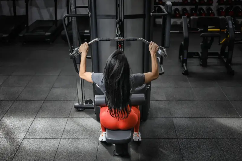 Mujer con cabello largo y negro realiza ejercicios de antebrazo en polea, sentada en una máquina de gimnasio.